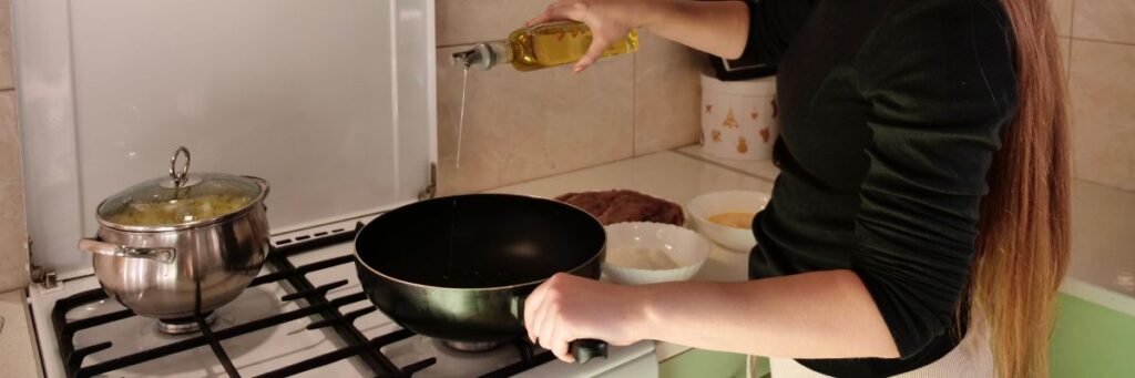 Woman pouring cooking oil into a pan on a kitchen stove, illustrating the use of cooking oils in daily meal preparation.