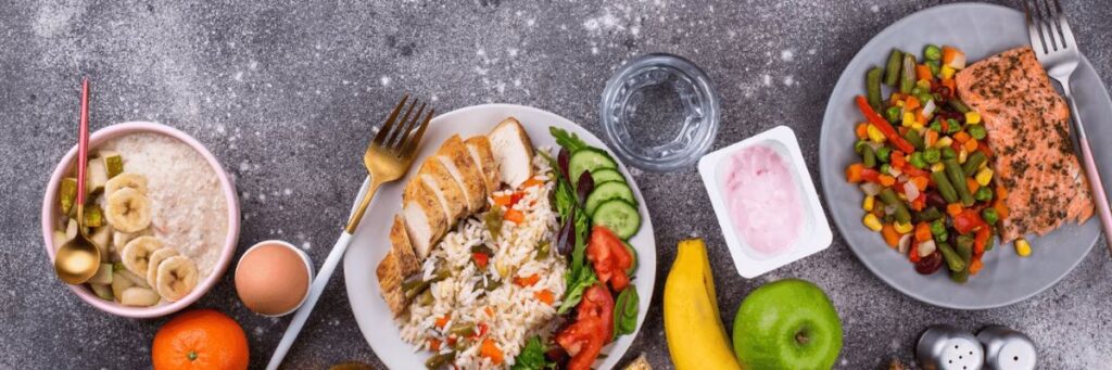 Balanced meal setup with rice, grilled chicken, vegetables, oatmeal, fruits, and fish on a gray table.
