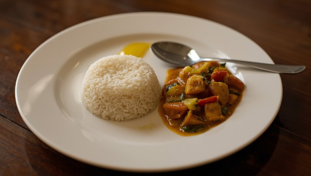 An image of a clean, home-cooked Indian dinner plate featuring a small serving of steamed white rice alongside dal, sautéed vegetables, and a spoonful of ghee. The plate is placed on a wooden dining table with soft warm evening lighting to suggest dinnertime.