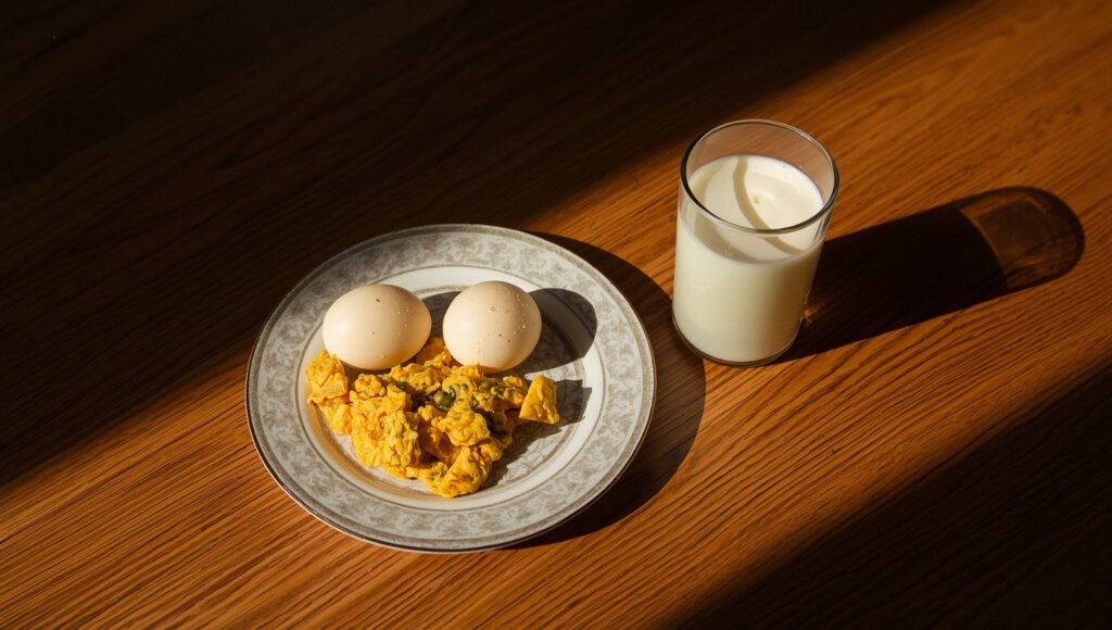 An image showing a healthy breakfast setup on a wooden table — a plate with 2 boiled or scrambled eggs next to a clear glass of milk. The lighting is warm and natural, suggesting a balanced and nutritious start to the day.
