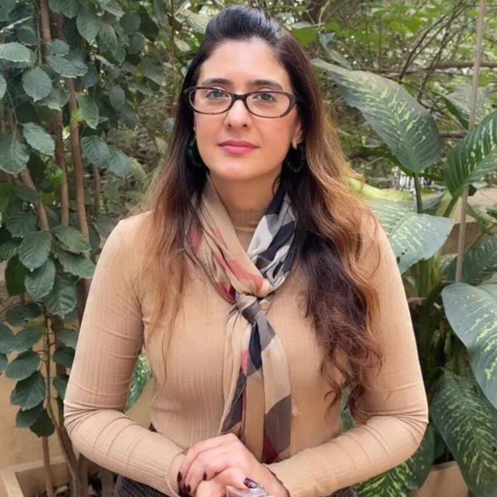 Pooja Makhija, famous dietitian in India, wearing beige top with patterned scarf and glasses, standing confidently outdoors amidst green plants, showcasing nutrition expertise.