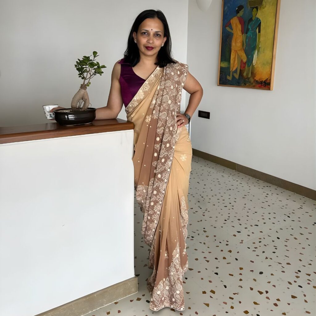 Rujuta Diwekar, famous dietitian in India, wearing a beige embroidered saree with purple blouse, standing elegantly indoors next to a bonsai plant and artwork, embodying Indian nutrition expertise