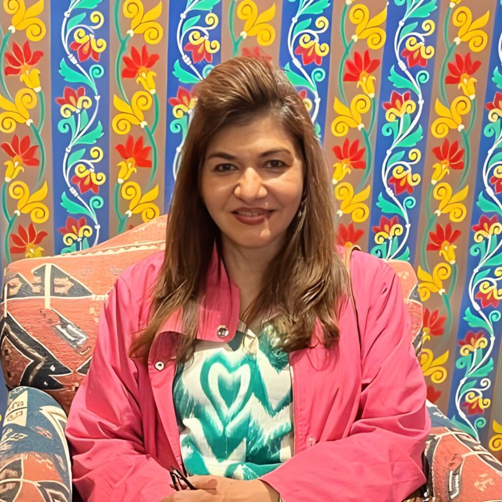 Anjali Mukherjee, famous dietitian in India, smiling warmly in a pink jacket over printed top, sitting against a colorful floral-patterned background, highlighting nutrition and wellness.
