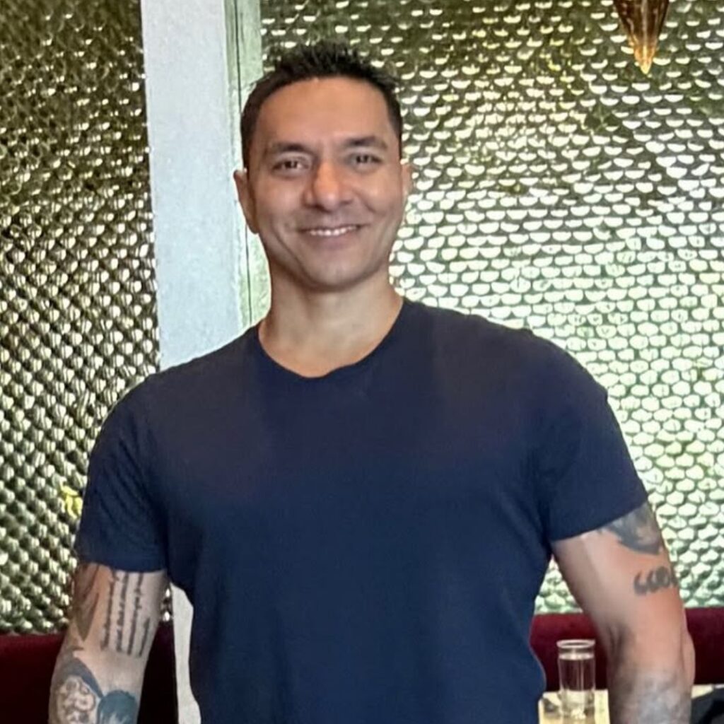 Luke Coutinho, famous dietitian in India, smiling confidently in a navy t-shirt with visible tattoos, standing against a golden textured wall, embodying holistic nutrition and wellness.